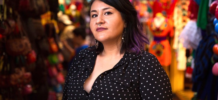 Chef Denice Vallejo is wearing a black shirt with shirt dark hair and mauve lipstick behind her a bustling market scene out of focus