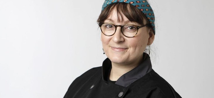 Kristina stands in a black chefs coat with a blue printed headband above her brown bangs. She has on dark cat eye framed glasses and hanging earrings