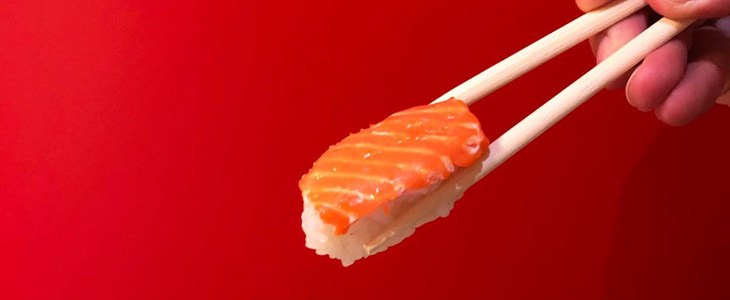 Credit:VegNews plant-based salmon sushi between two chopsticks on a red background