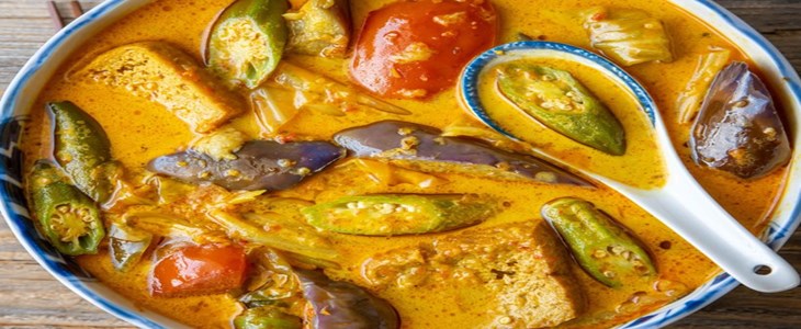 On a wood grain table sits a white bowl filled with bright yellow broth and summer vegetables