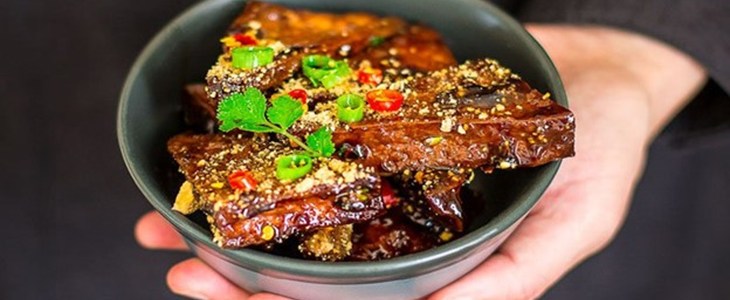 Hand holding black bowl of braised tofu in black room