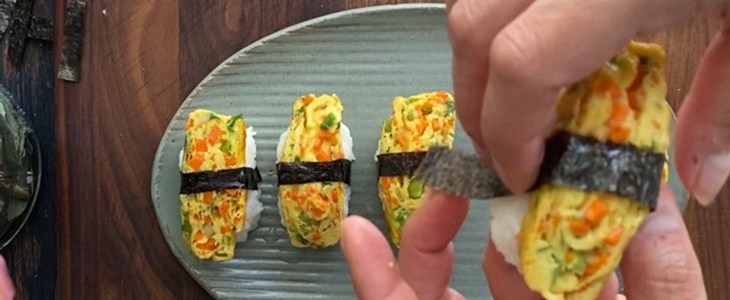 On a dark wood table sits a grey plate with three pieces of sushi sittig horizontally on a plate. A hand holds one piece of sushi in the foreground