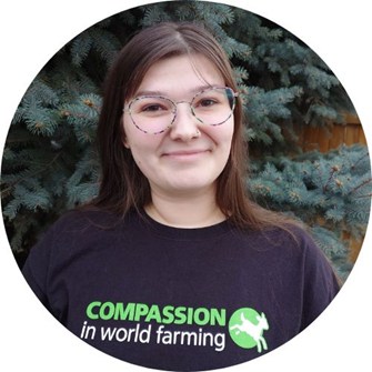 Anna smiling at camera in front of a pine tree wearing glasses and her Compassion t-shirt.
