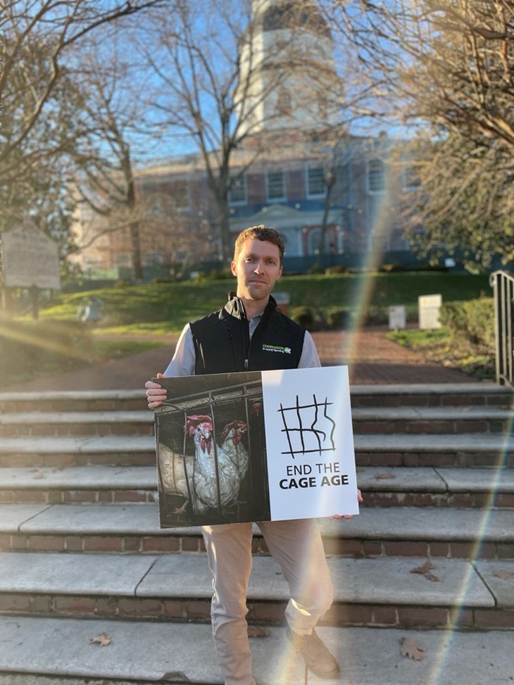 U.S. Staff Member Ryan Robinson stands on the steps in front of a government building, holding a sign showing a caged hen that says "End the Cage Age."